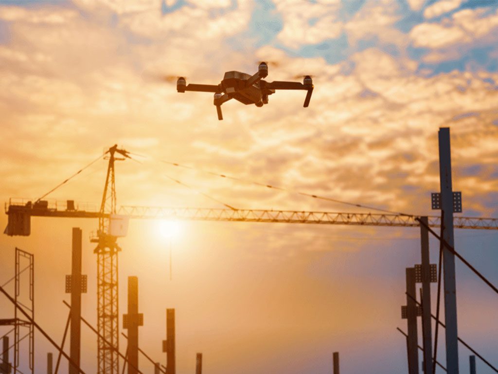 Drone flying over a construction site at sunset, silhouetted against cranes and steel structures.