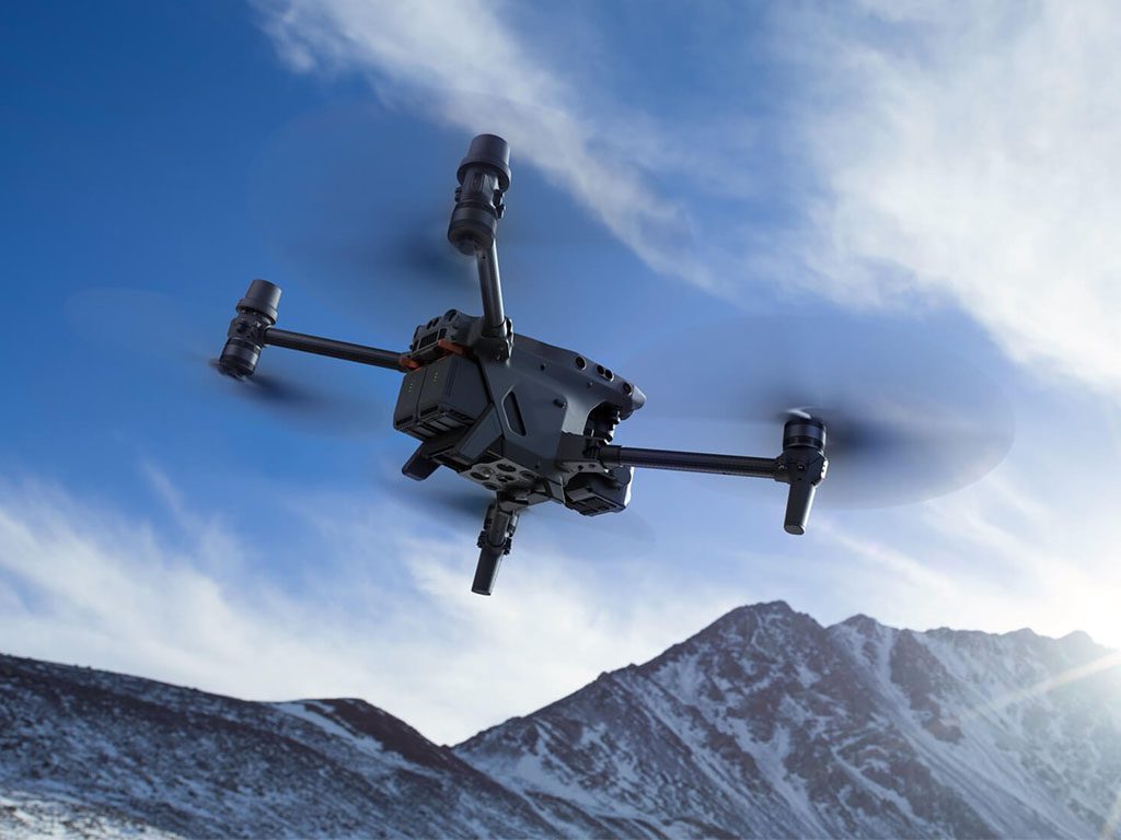 Drone DJI Matrice 30T en vol devant des montagnes enneigées sous un ciel bleu.