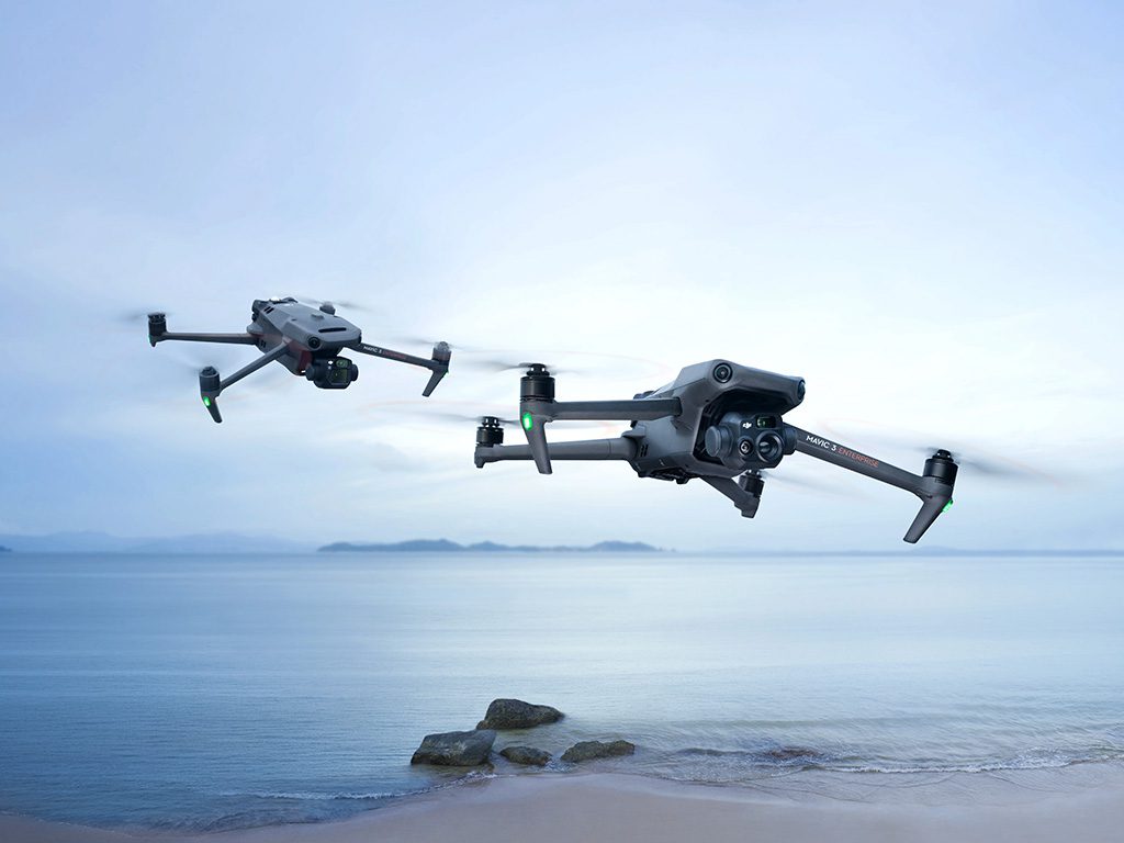Deux drones DJI Mavic survolant une plage paisible au crépuscule, avec vue sur une mer calme et des îles lointaines.